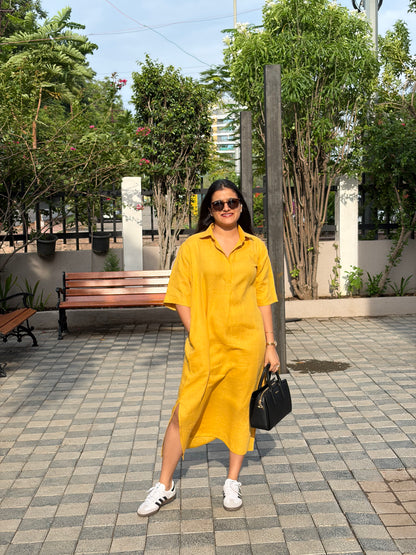 Woman in a yellow linen loose fit dress standing on a paved area with greenery in the background