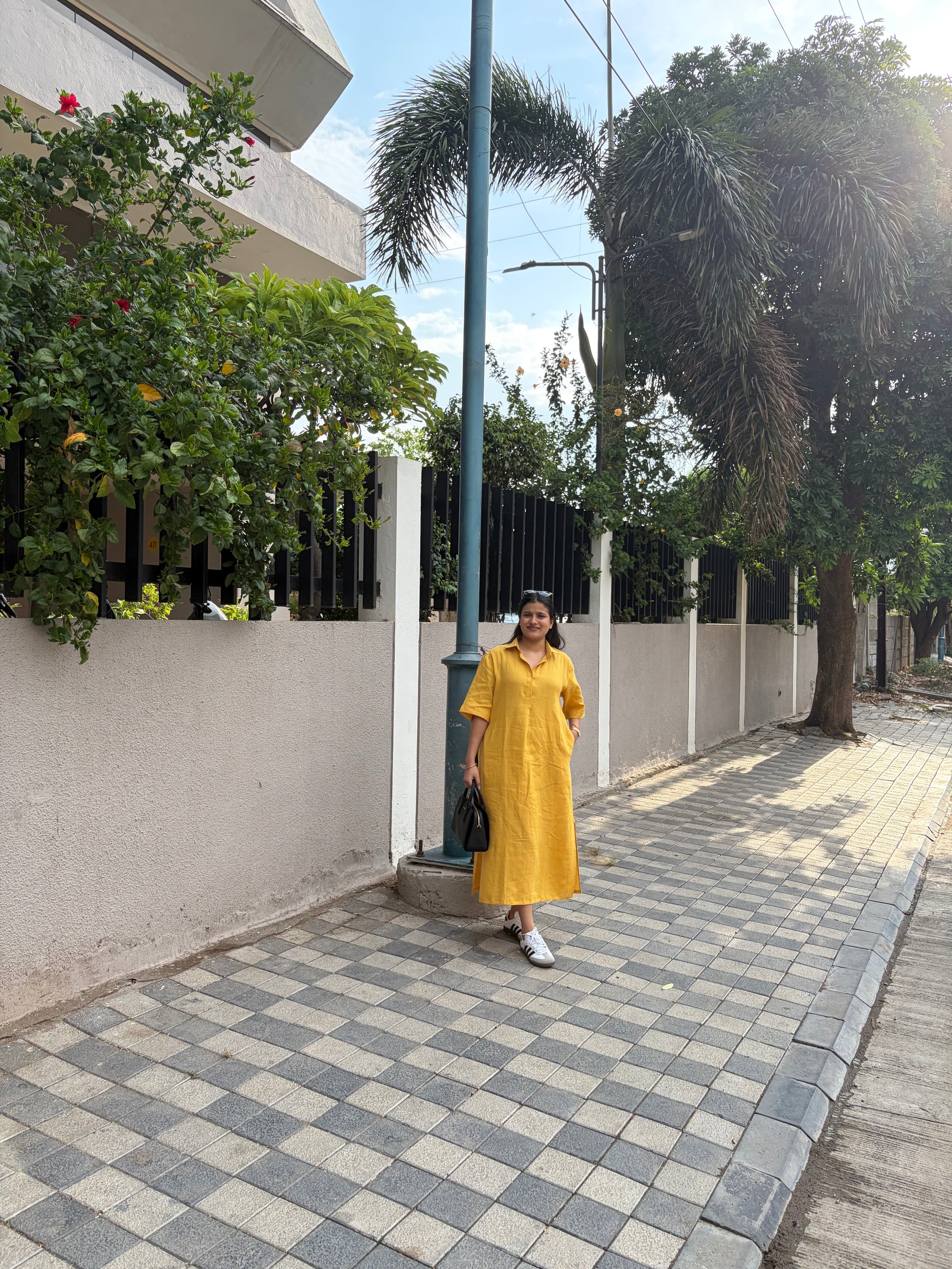 Woman in a yellow dress walking on a sidewalk with greenery and buildings in the background
