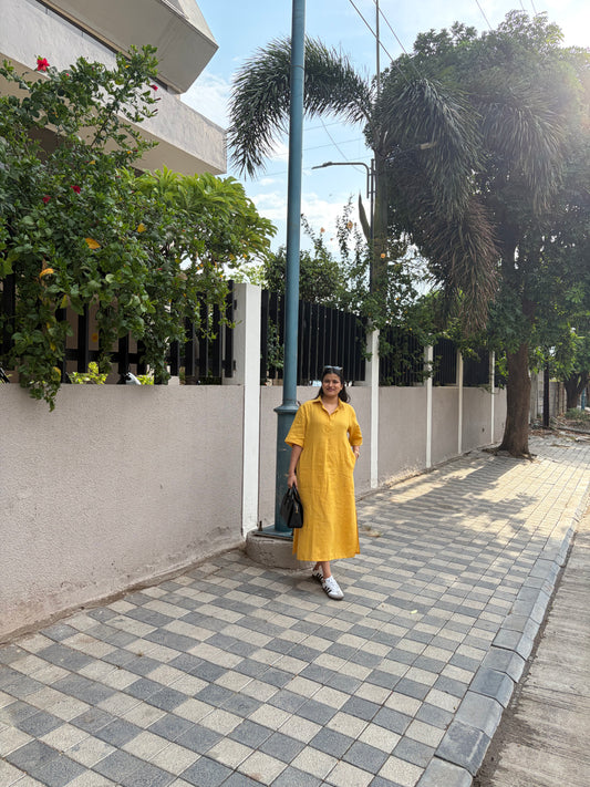 Woman in a yellow dress walking on a sidewalk with greenery and buildings in the background