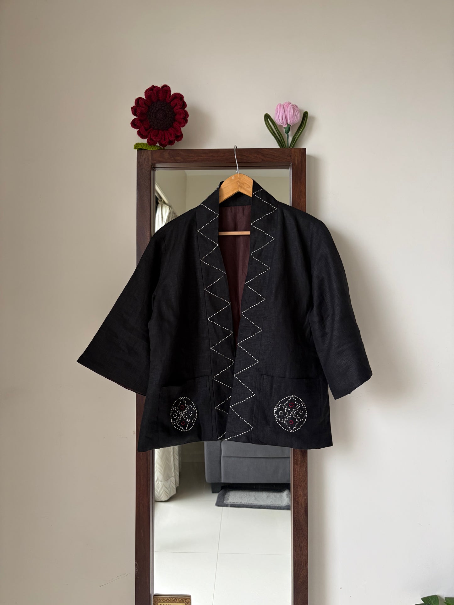 A black Japanese-style jacket with white zigzag and circular embroidery, hanging on a wooden hanger in front of a mirror.