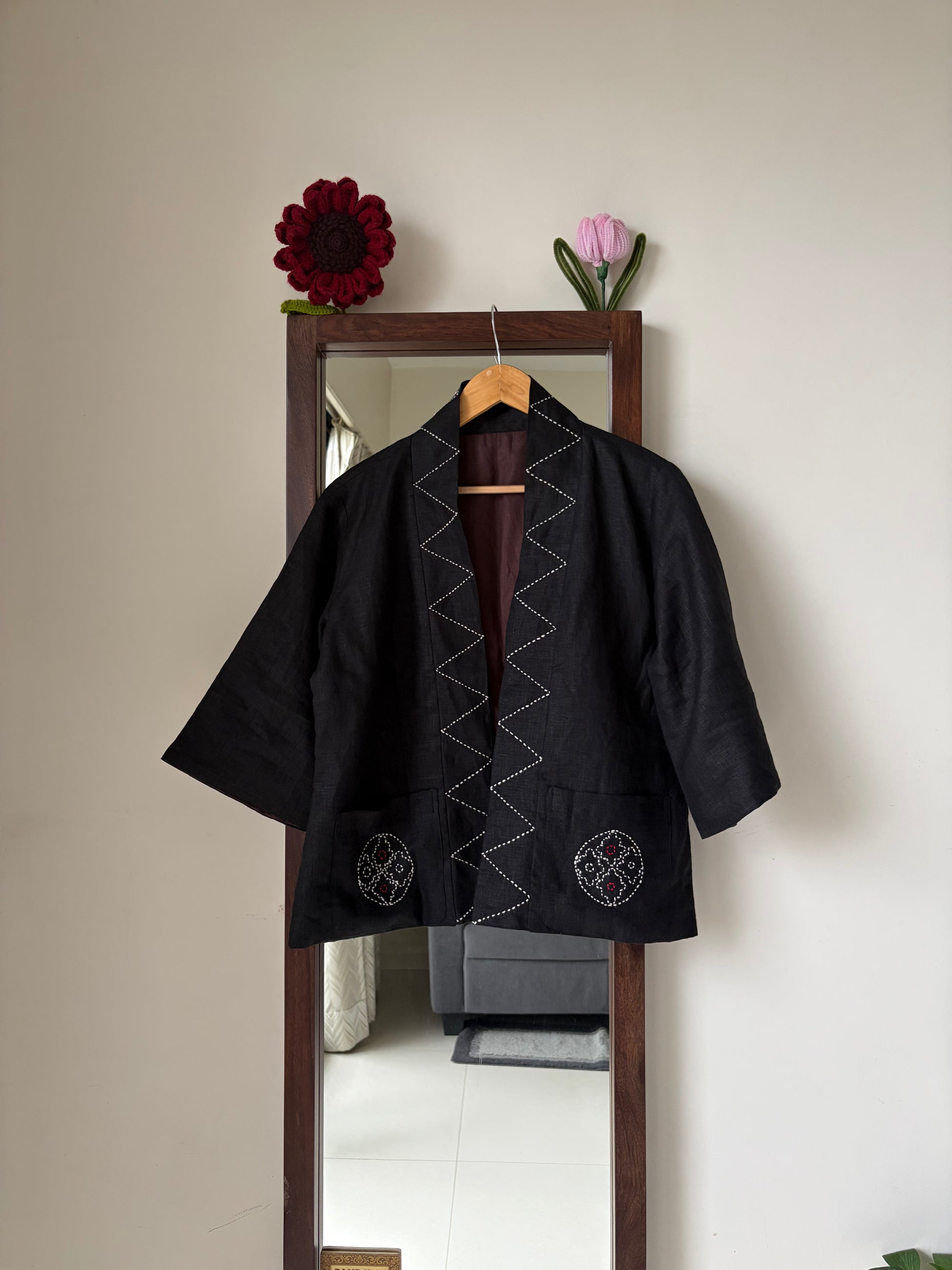 A black Japanese-style jacket with white zigzag and circular embroidery, hanging on a wooden hanger in front of a mirror.