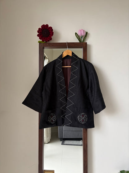 A black Japanese-style jacket with white zigzag and circular embroidery, hanging on a wooden hanger in front of a mirror.
