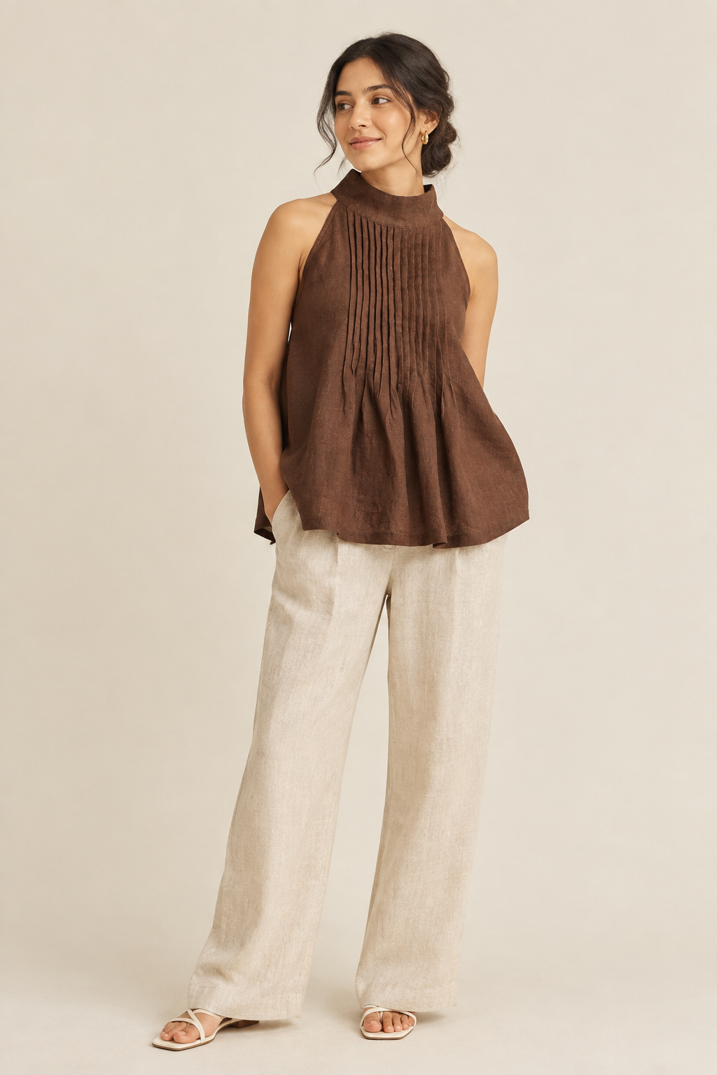 A woman modeling a sleeveless brown linen halter top with vertical pleating and a flared peplum hem, paired with wide-leg cream pants and flat sandals, against a plain beige studio background.
